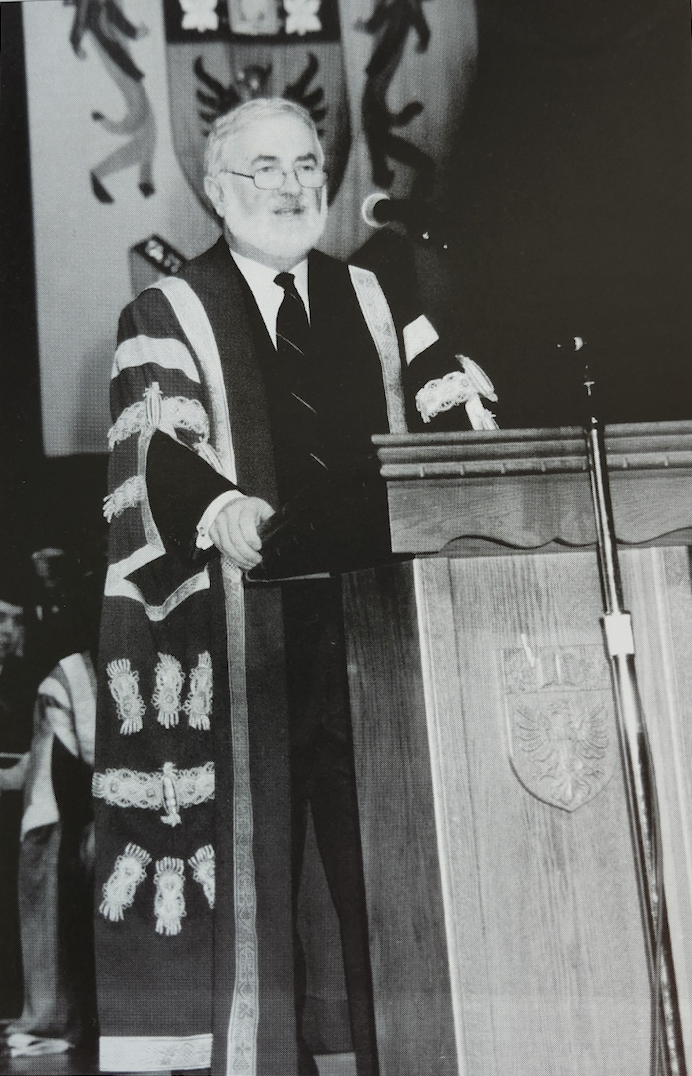 A black and white photo of a man in ceremonial robes, speaking at a lectern. 